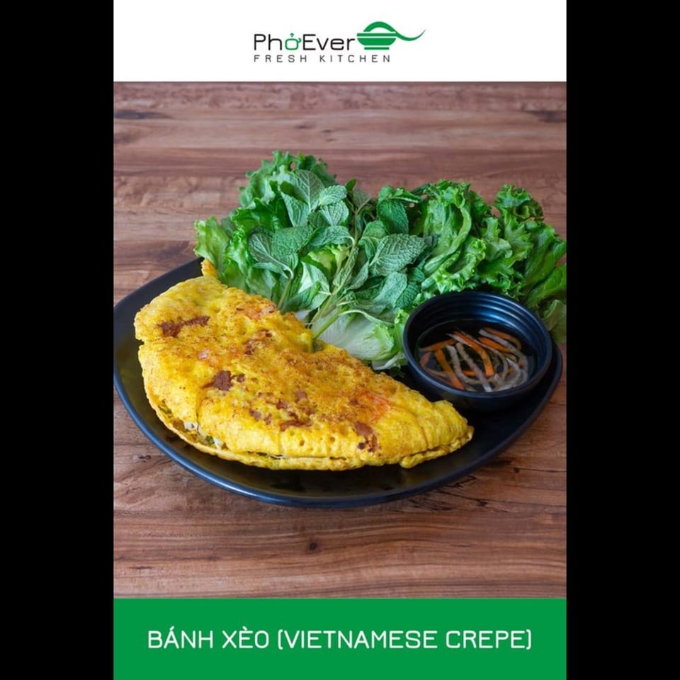 A golden Vietnamese crepe (Bánh Xèo) with fresh lettuce, mint, and dipping sauce on a wooden table.