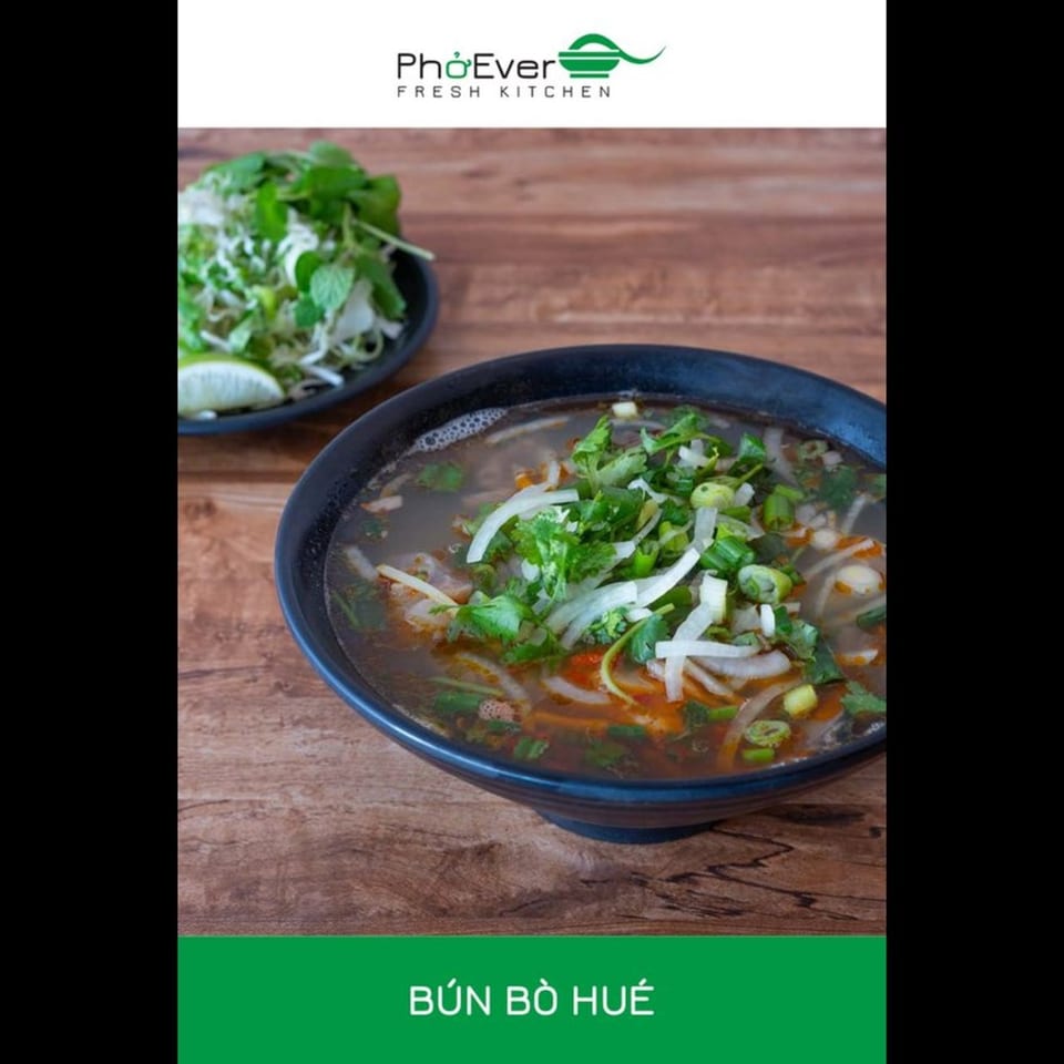A bowl of PhoEver Bún Bò Huế soup with herbs, onions, and lime on a wooden table.
