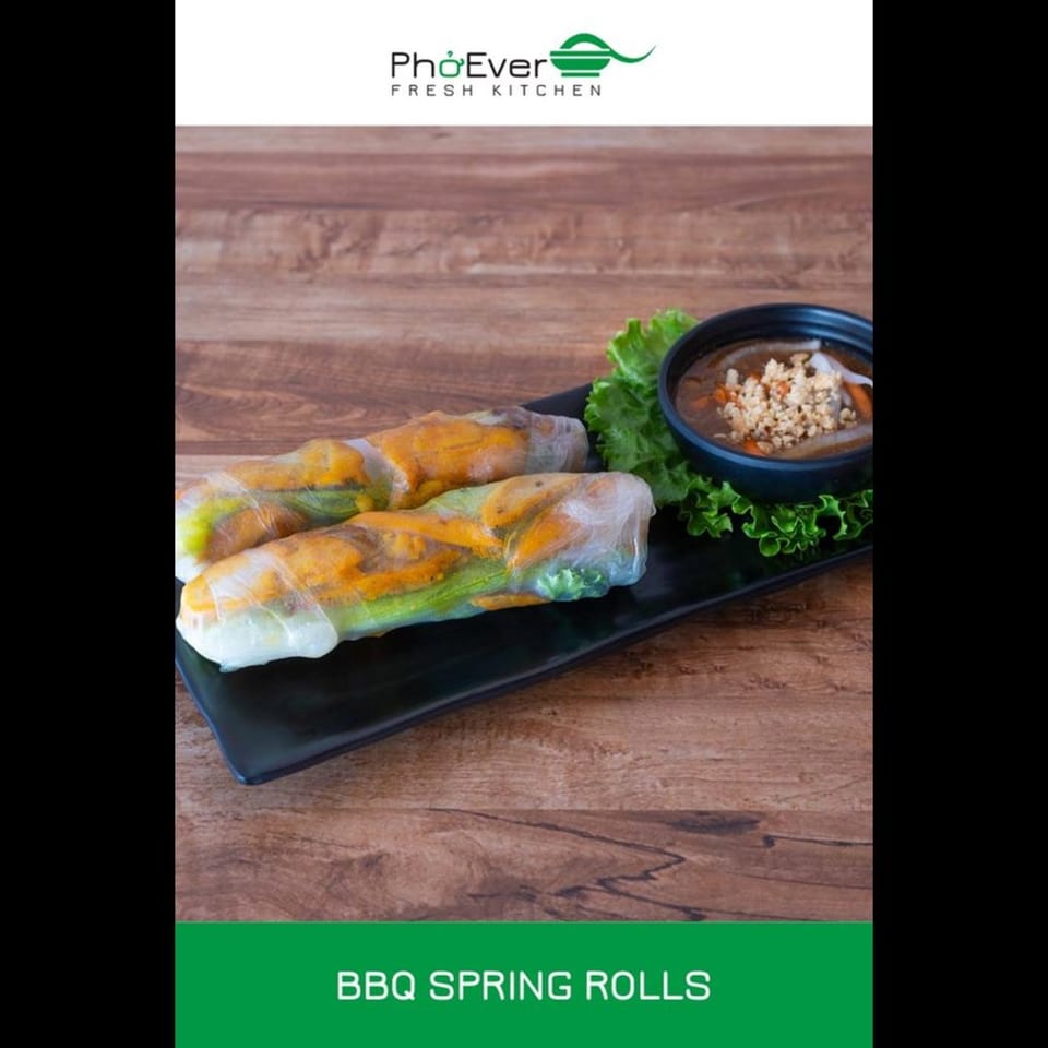 Two BBQ spring rolls with lettuce and dipping sauce on a black plate, on a wooden table.