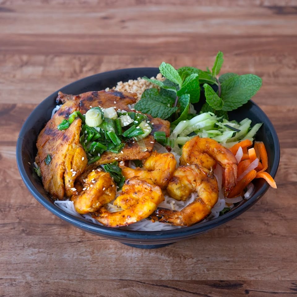 A Vietnamese vermicelli bowl with grilled pork, shrimp, fresh herbs, and vegetables on a wooden table.