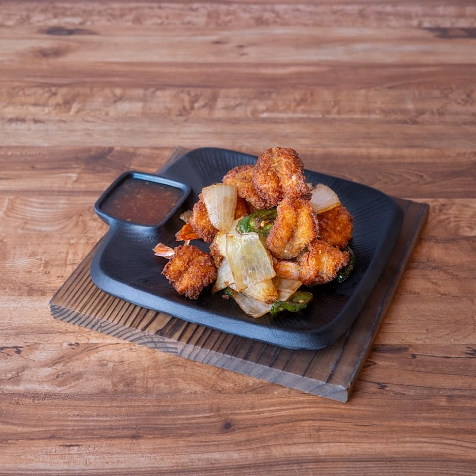Crispy fried shrimp with sautéed onions and jalapeños, served with a dipping sauce on a black plate.