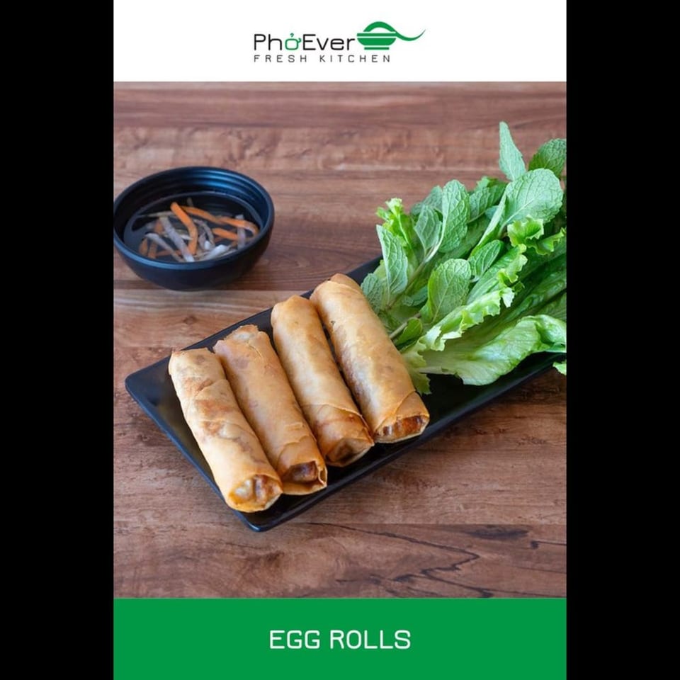 Four crispy egg rolls on a black plate with lettuce, mint, and a small bowl of sauce.