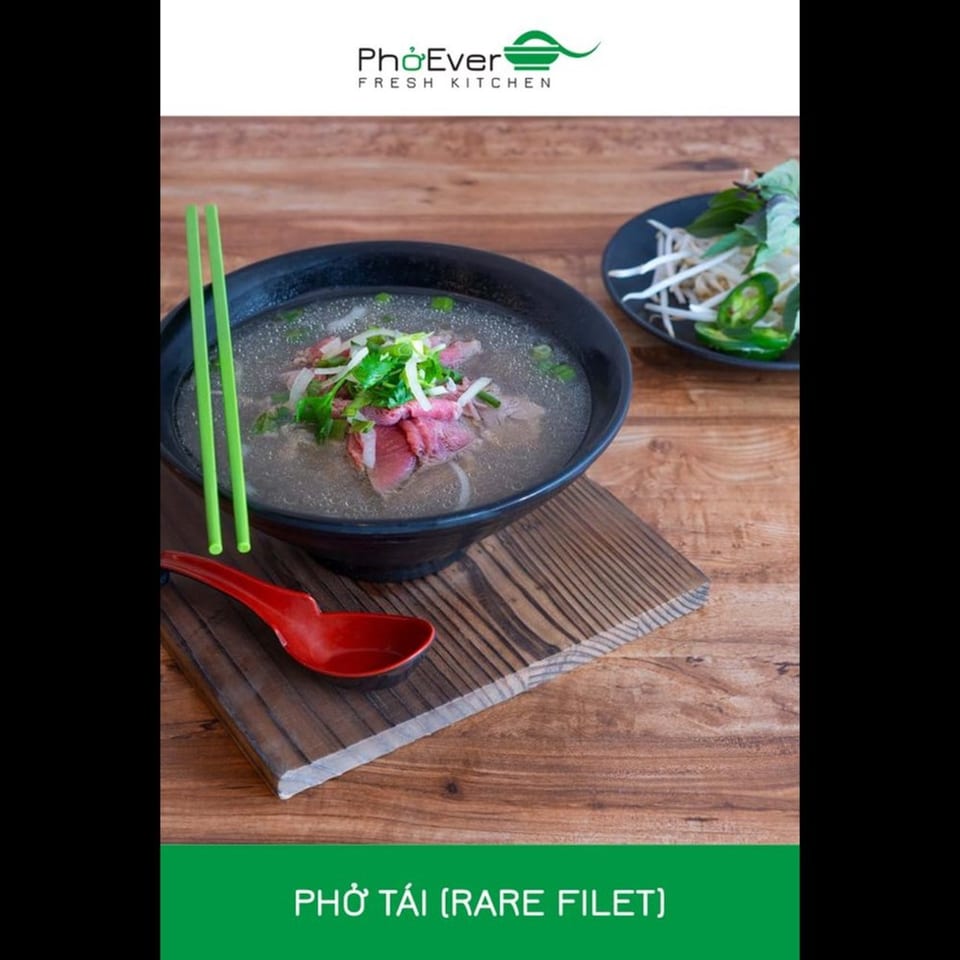 A bowl of Pho Tai (Rare Filet) with green chopsticks and a red spoon on a wooden table.