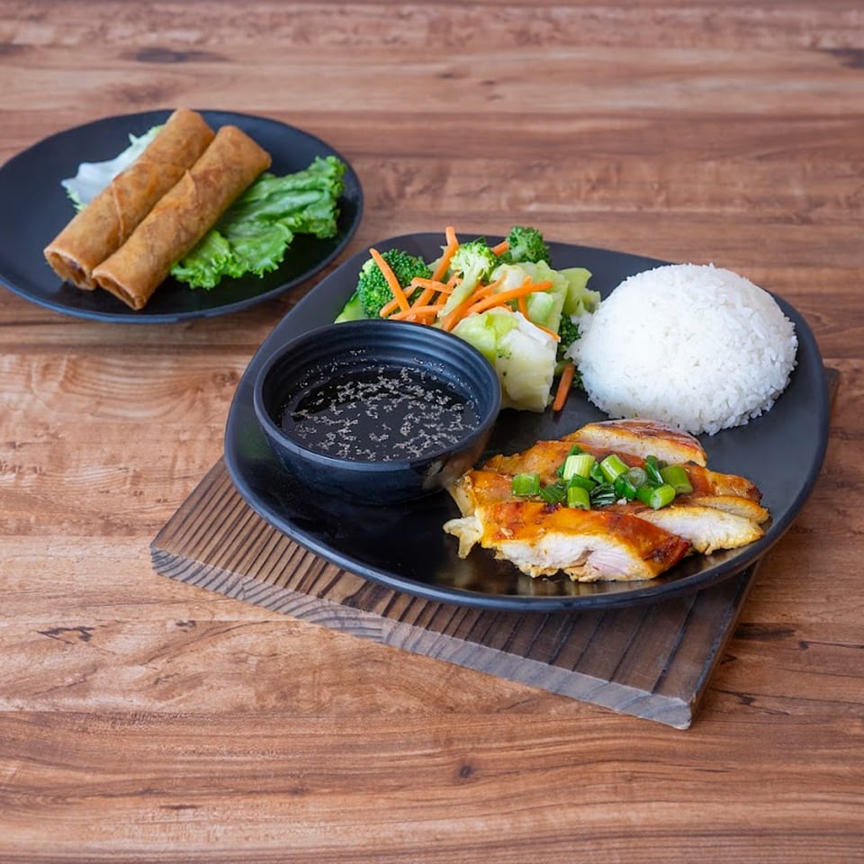 Vietnamese grilled chicken with rice, steamed vegetables, dipping sauce, and two spring rolls.